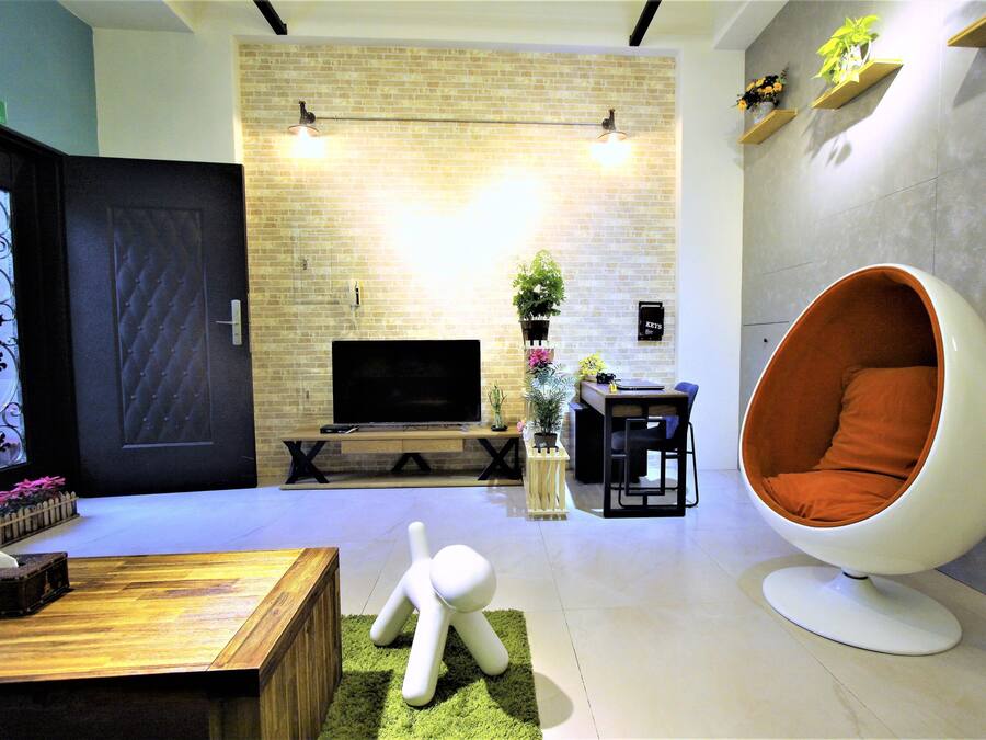 A modern living room with a brick wall, a flat-screen TV, a wooden coffee table, a unique orange chair, and a small table with a plant.