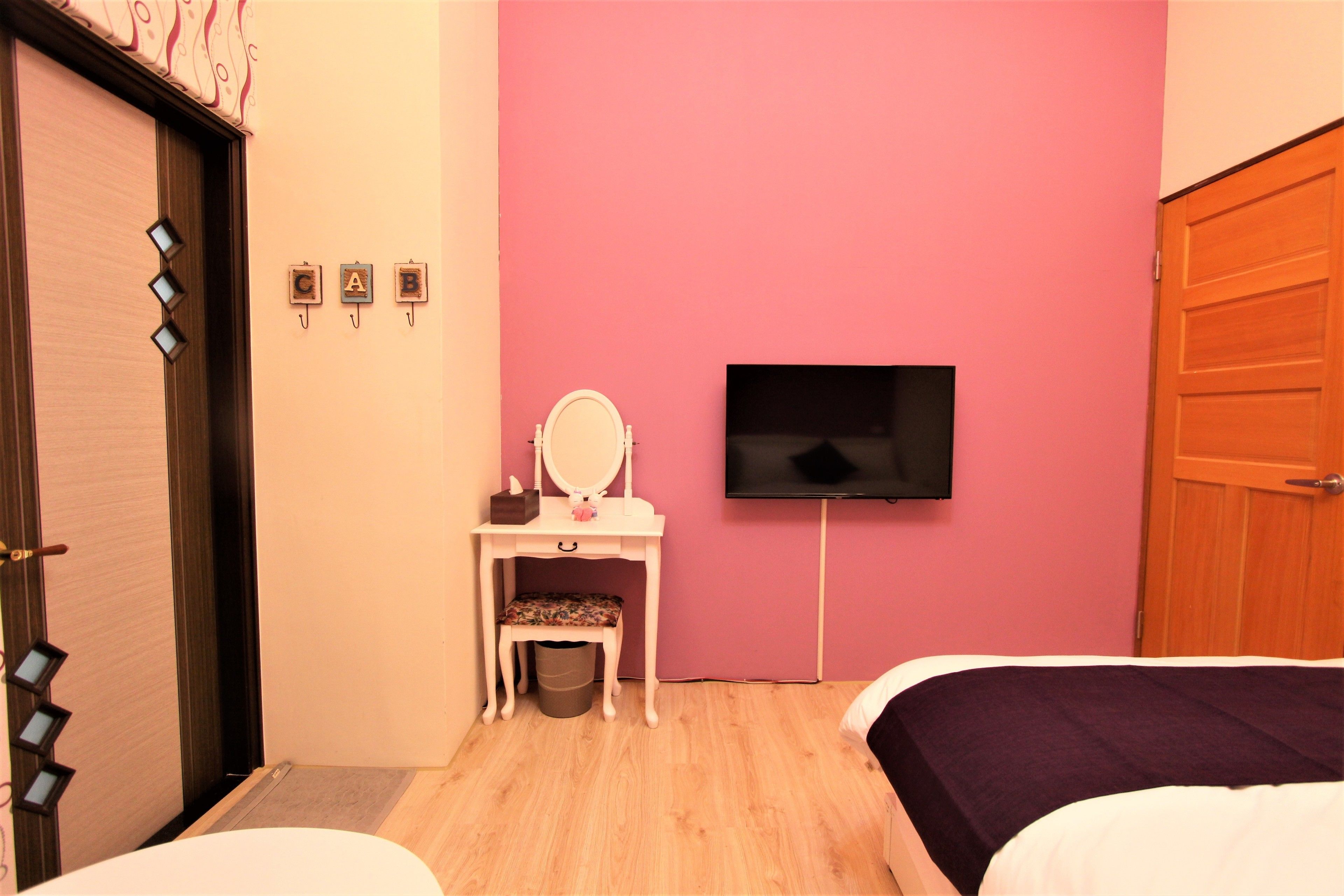 A room with a pink wall, a wooden door, a desk with a mirror, a television, and two beds.
