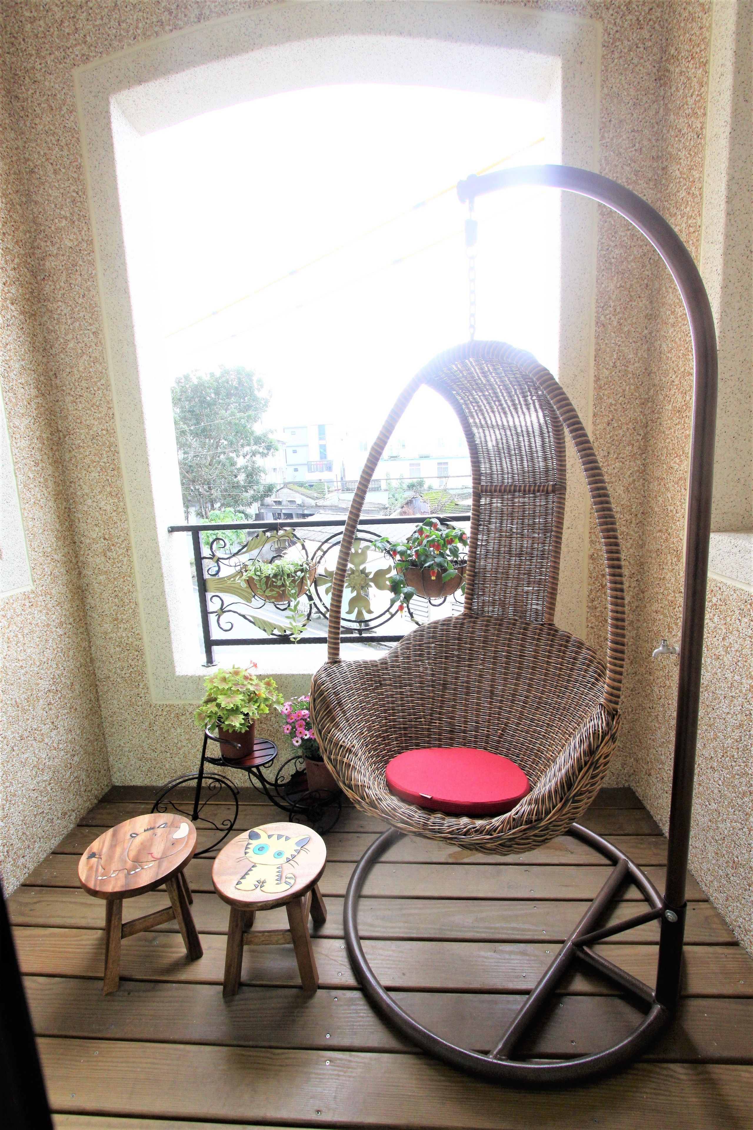 A wicker swing chair with a red cushion, two small wooden stools, and a potted plant on a balcony with a view of buildings and trees.