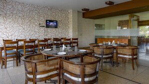 A restaurant with round tables and wicker chairs, a TV mounted on the wall, and a patterned wall. - Shivam Resorts (Jaipur)
