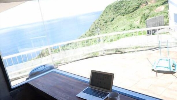 A laptop on a wooden table by a window with a view of a green hillside and the sea. - Happy coral (Higashi)