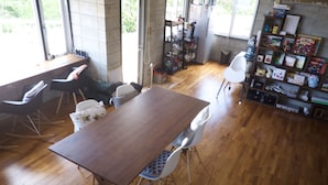 A dining area with a wooden table and chairs, a bookshelf with various items, and a window providing natural light. - Happy coral (Higashi)