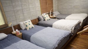 A hotel room with three beds arranged in a row, each with a pillow and a blanket. There is a wooden floor and a window with blinds. - Happy coral (Higashi)