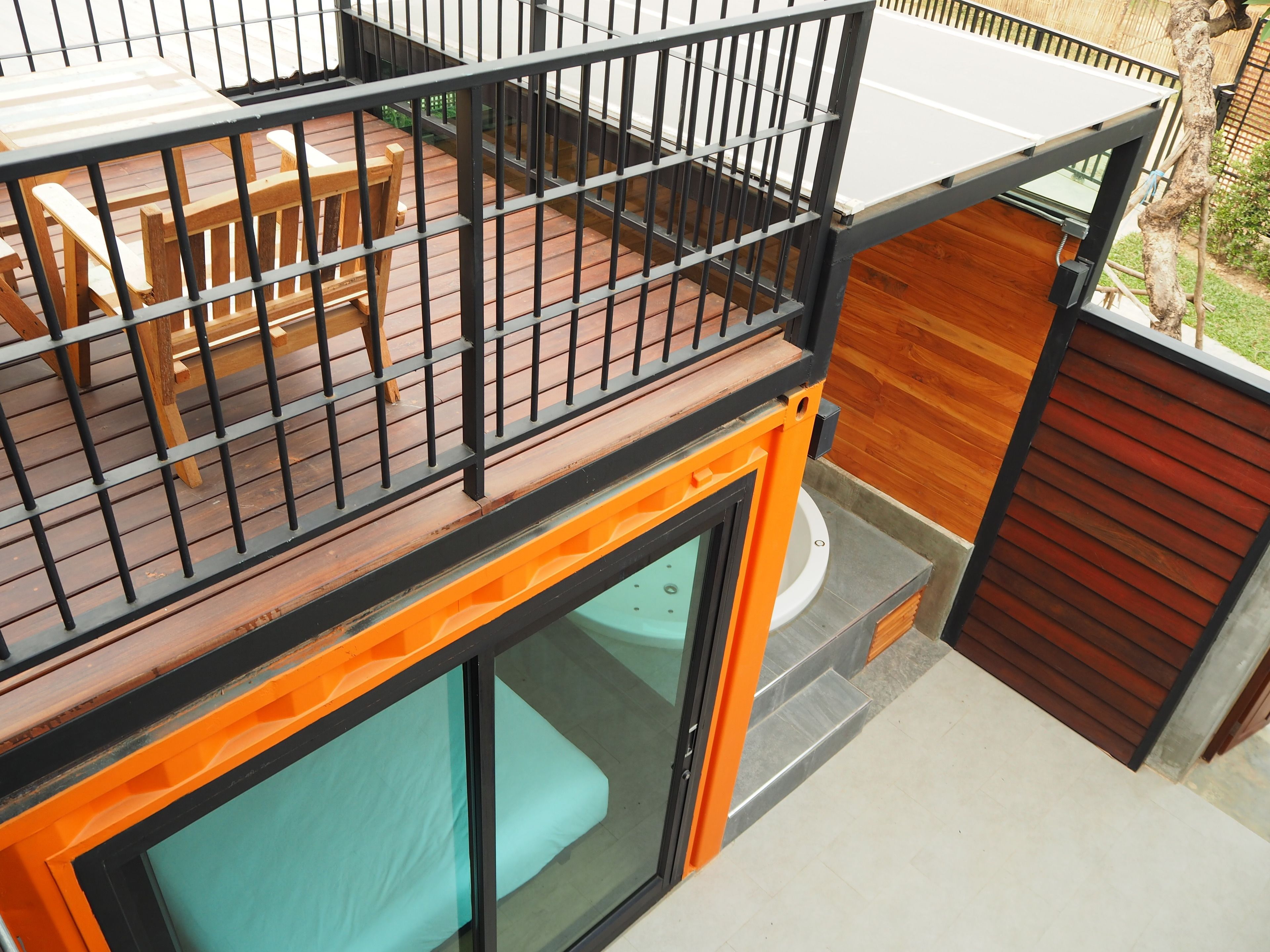 A modern outdoor deck with a wooden table and chairs, a glass door leading to a patio area, and a view of a garden.