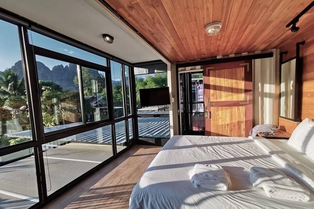 Une chambre avec un grand lit, un plafond en bois et une vue sur les montagnes, visible à travers de grandes fenêtres.