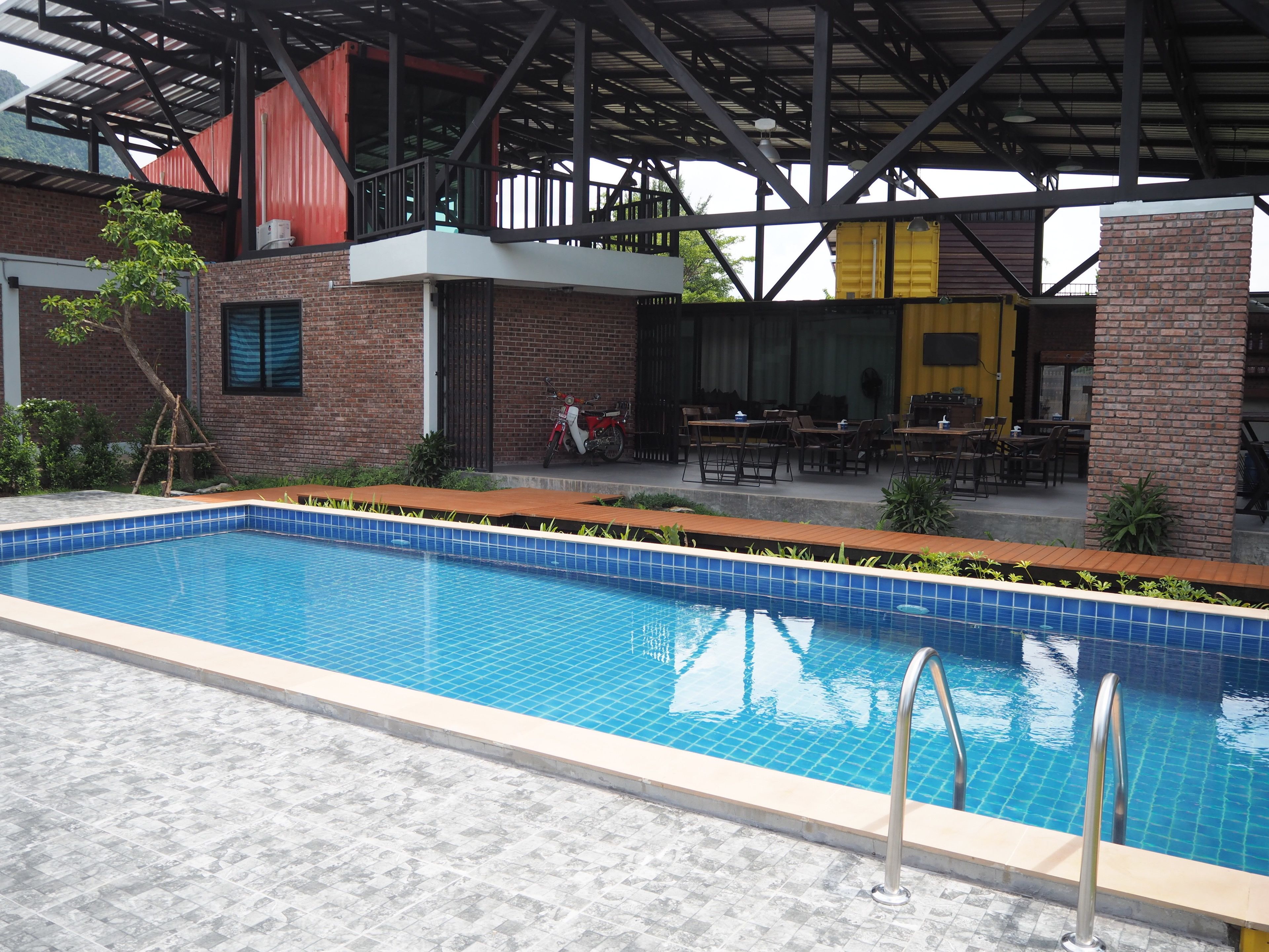 A swimming pool with a tiled edge, surrounded by a patio area with outdoor seating and a brick building in the background.