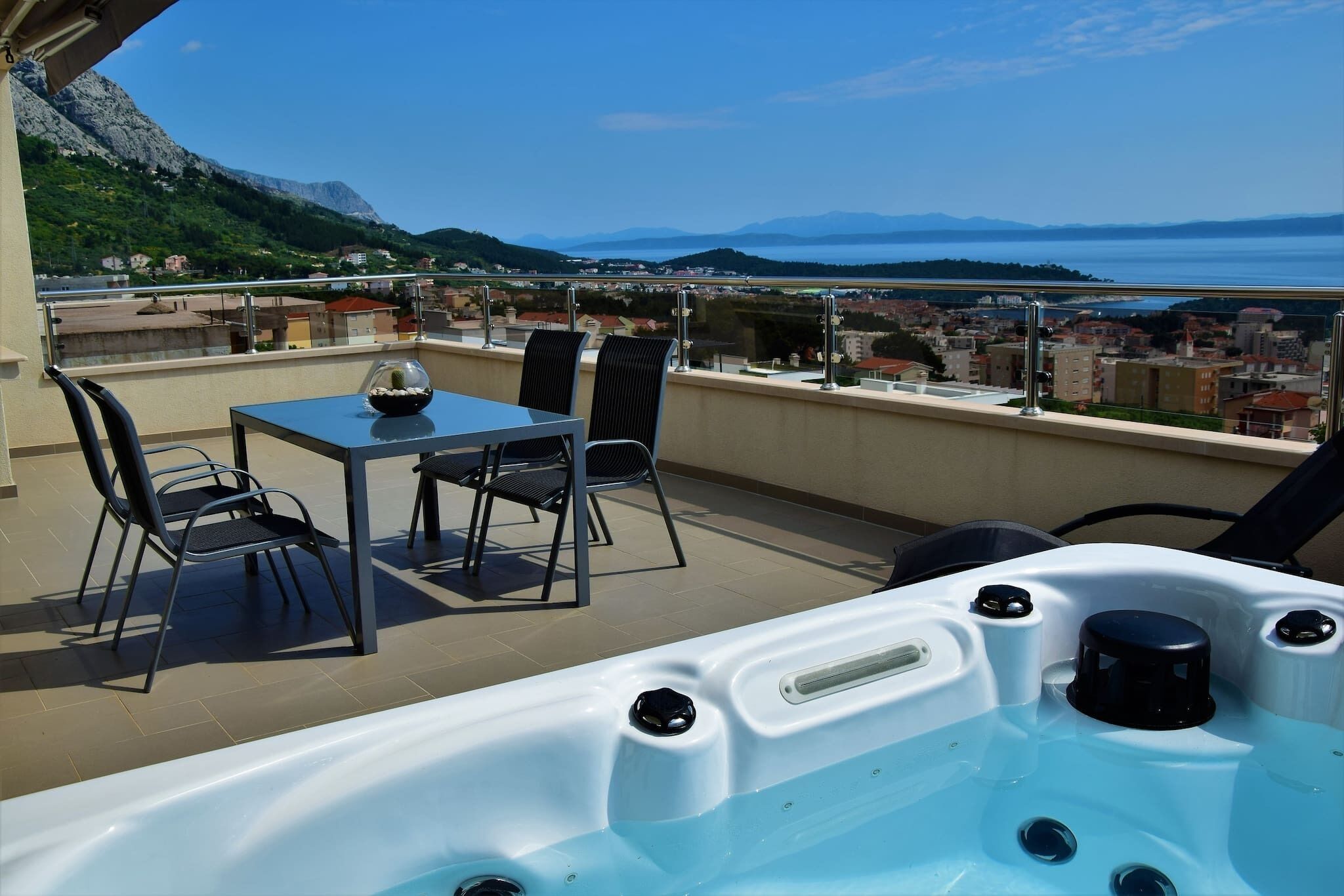 A balcony with a hot tub, a table, and chairs, overlooking a scenic view of a lake and mountains.