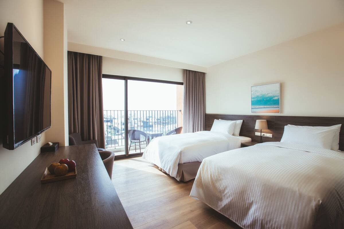 Ein Hotelzimmer mit zwei Betten, einem Fernseher, einem Schreibtisch mit Früchten und einem Balkon mit Blick auf die Stadt.
