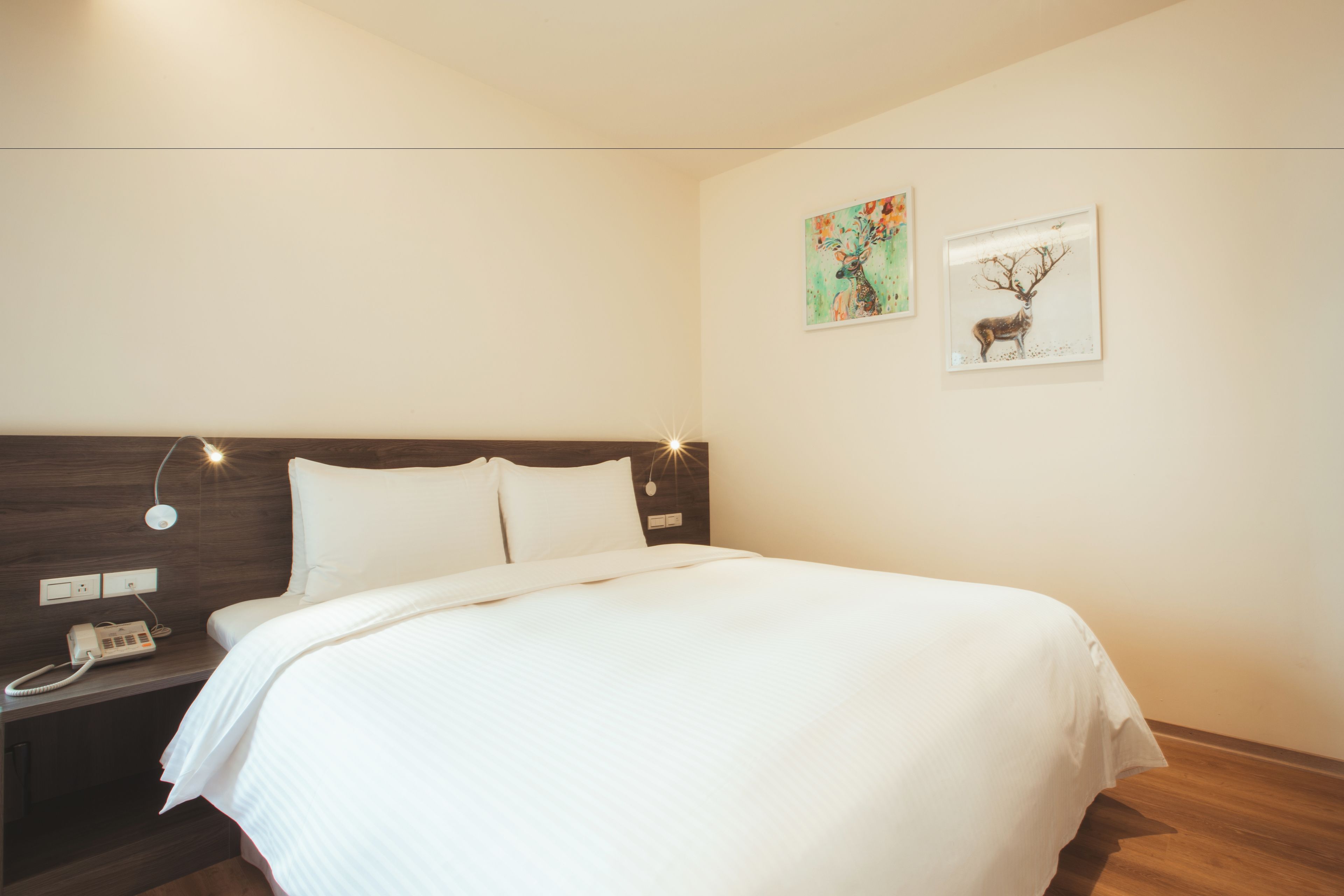 Una cama bien hecha en una habitación de hotel con cabecera de madera, un teléfono en la mesita de noche y dos cuadros en la pared.