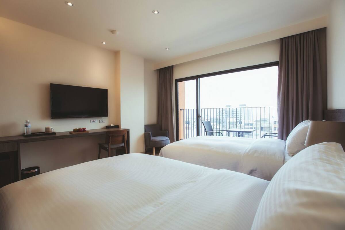 Ein Hotelzimmer mit zwei Betten, einem Schreibtisch, einem Stuhl, einem Fernseher und einem Balkon mit Blick auf die Stadt.