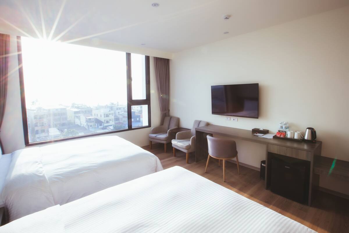 Ein Hotelzimmer mit zwei Betten, einem Schreibtisch, einem Sessel und einem Fernseher, mit Blick auf die Stadt.
