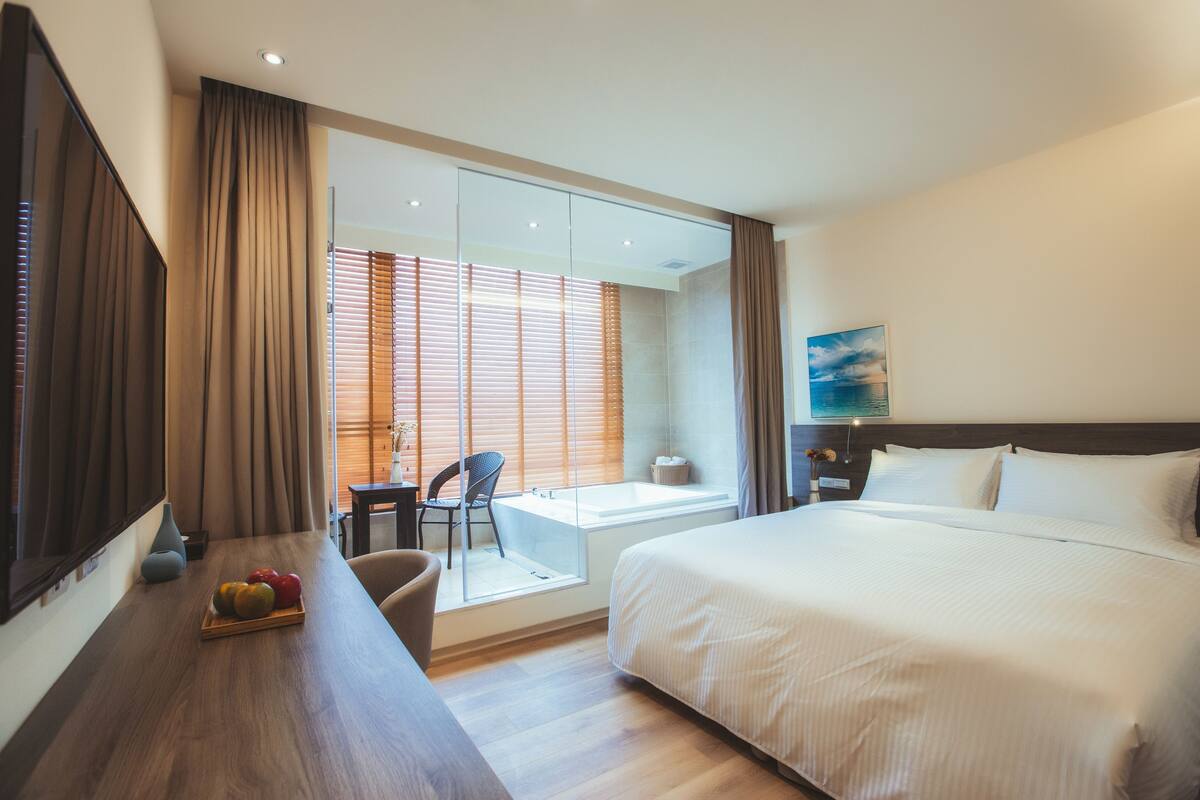 Ein Hotelzimmer mit einem großen Bett, einem Schreibtisch mit Obstteller, einem Sessel, einem Fenster mit Jalousien und einem Gemälde an der Wand.