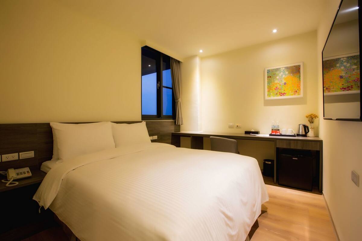 Ein Hotelzimmer mit Bett, Schreibtisch, Stuhl, Telefon, Fenster und einem Bild an der Wand.