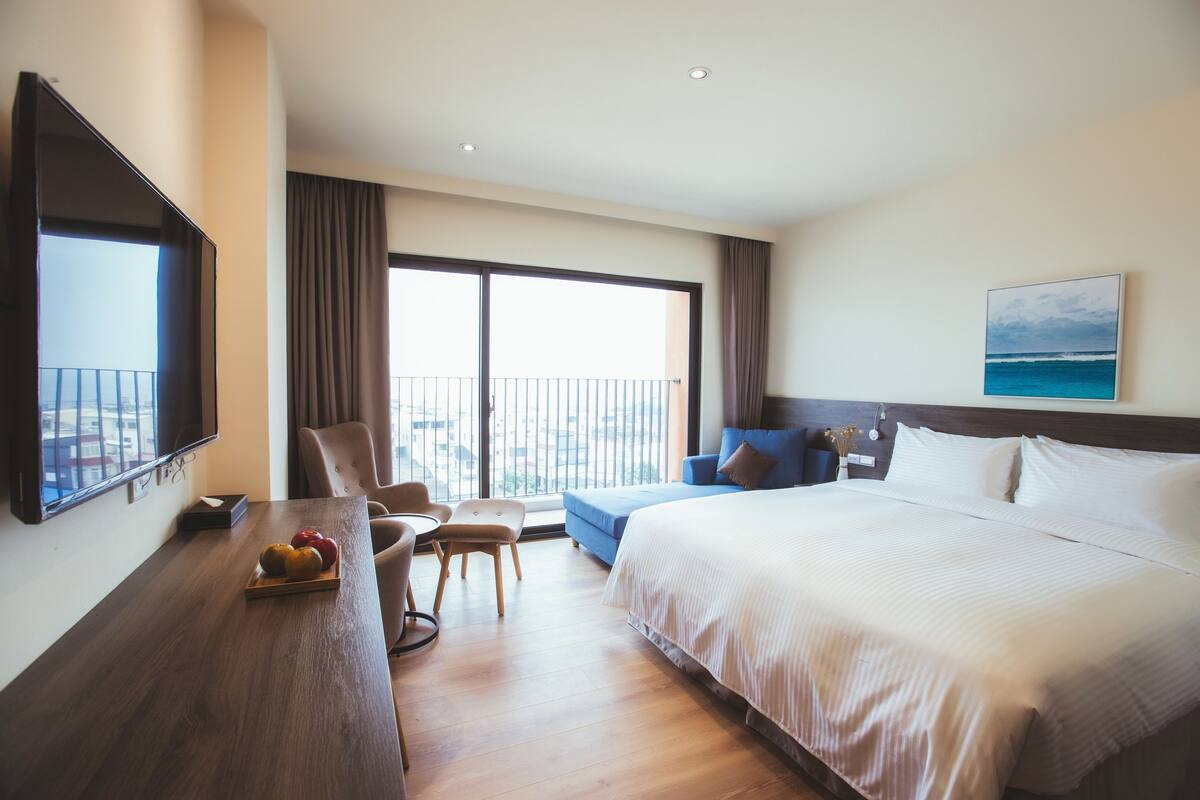 Ein Hotelzimmer mit einem großen Bett, einem Schreibtisch mit Obstschale, einem Sessel, einer Couch und einem Balkon mit Blick.
