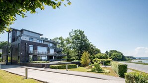 Un bâtiment moderne à deux étages, doté de grandes fenêtres et d’un balcon, entouré d’un espace verdoyant bien entretenu et offrant une vue dégagée sur la mer.