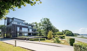 A modern two-story building with large windows and a balcony, surrounded by well-maintained greenery and a clear view of the sea. - Exclusive apartment with water view - Apartment Meierwik (Glücksburg (Ostsee))