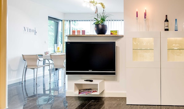 A modern living room with a flat-screen TV, a dining area with chairs, a potted plant, candles, and wine bottles. - Exclusive apartment with water view - Apartment Meierwik (Glücksburg (Ostsee))