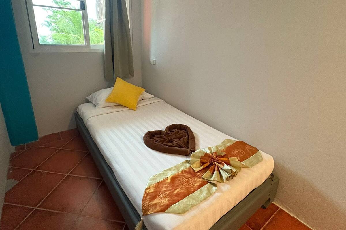 A single bed with a white mattress, a heart-shaped pillow, and a decorative bow on the bedspread. There is a window with a view of greenery outside, and an air conditioning unit mounted on the wall.