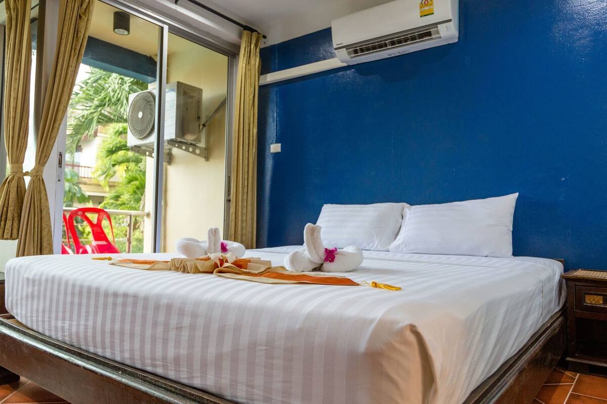 A hotel room with a bed, a red chair, an air conditioning unit, and a view of greenery outside.