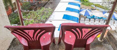 A balcony with two red plastic chairs, overlooking a hotel area with striped umbrellas and palm trees.