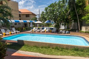A swimming pool with lounge chairs and umbrellas, surrounded by greenery and a building with a red roof. - Porterhouse Beach Hotel (Patong)