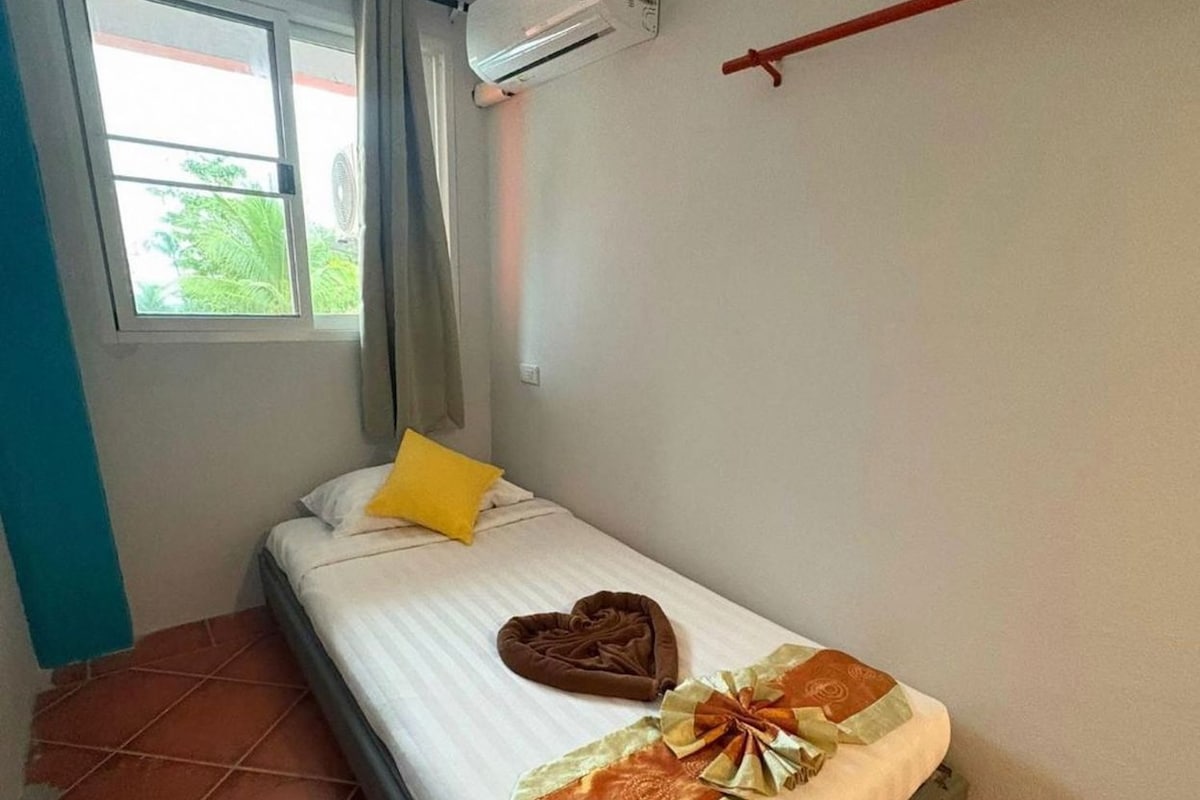 Quarto pequeno com uma cama de solteiro, janela, ar condicionado e uma cama com travesseiro em formato de coração e travesseiro decorativo.
