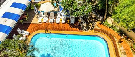 An aerial view of a swimming pool with lounge chairs and umbrellas, surrounded by greenery and a covered area with a striped awning.