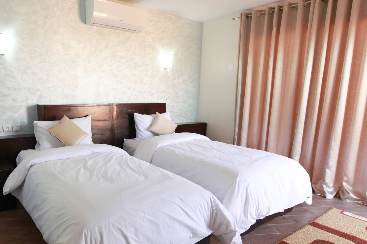 Habitación de hotel con dos camas individuales, cabecero de madera, aire acondicionado de pared y un ventanal con cortinas.
