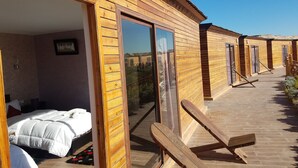 Una habitación tipo cabaña de madera con una cama, una mesita pequeña y un ventanal corredero que da a un espacio exterior con terraza de madera y sillas de descanso.
