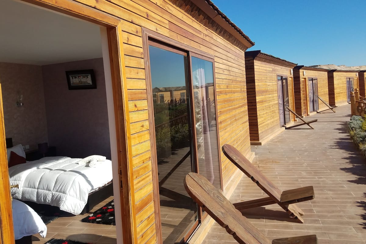Una habitación tipo cabaña de madera con una cama, una mesita pequeña y un ventanal corredero que da a un espacio exterior con terraza de madera y sillas de descanso.