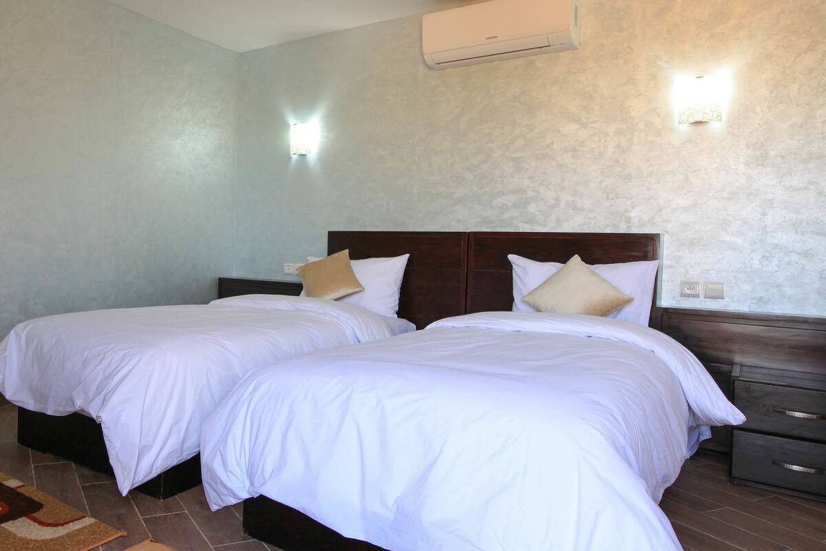Habitación de hotel con dos camas, cabecero de madera oscura, alfombra marrón y beige, y aire acondicionado instalado en la pared.