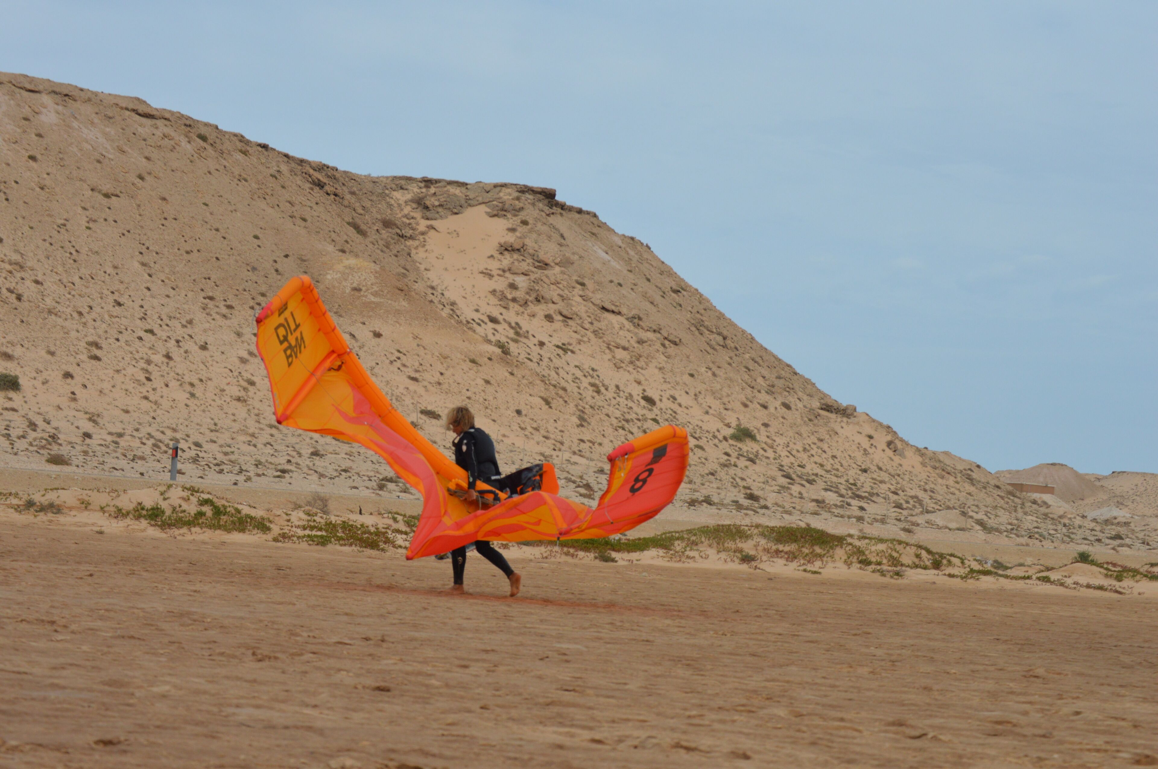 A person walking with a large orange kite in a desert landscape.