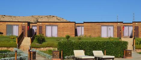 A row of wooden cabins with a staircase leading to the entrance, a small garden area with shrubs and a bench, and a clear blue sky.
