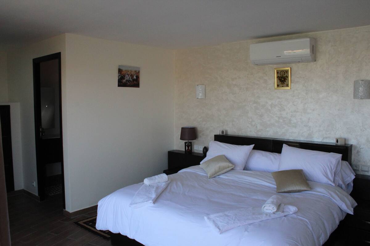 Una cama bien tendida con sábanas blancas, una mesita de noche con lámpara y un aire acondicionado de pared.