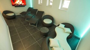 Ett modernt vardagsrum med en platt-TV som visar en bild av en öppen spis, flera olika sittplatser, inklusive en soffa och fåtöljer, samt små fönster som släpper in naturligt ljus.