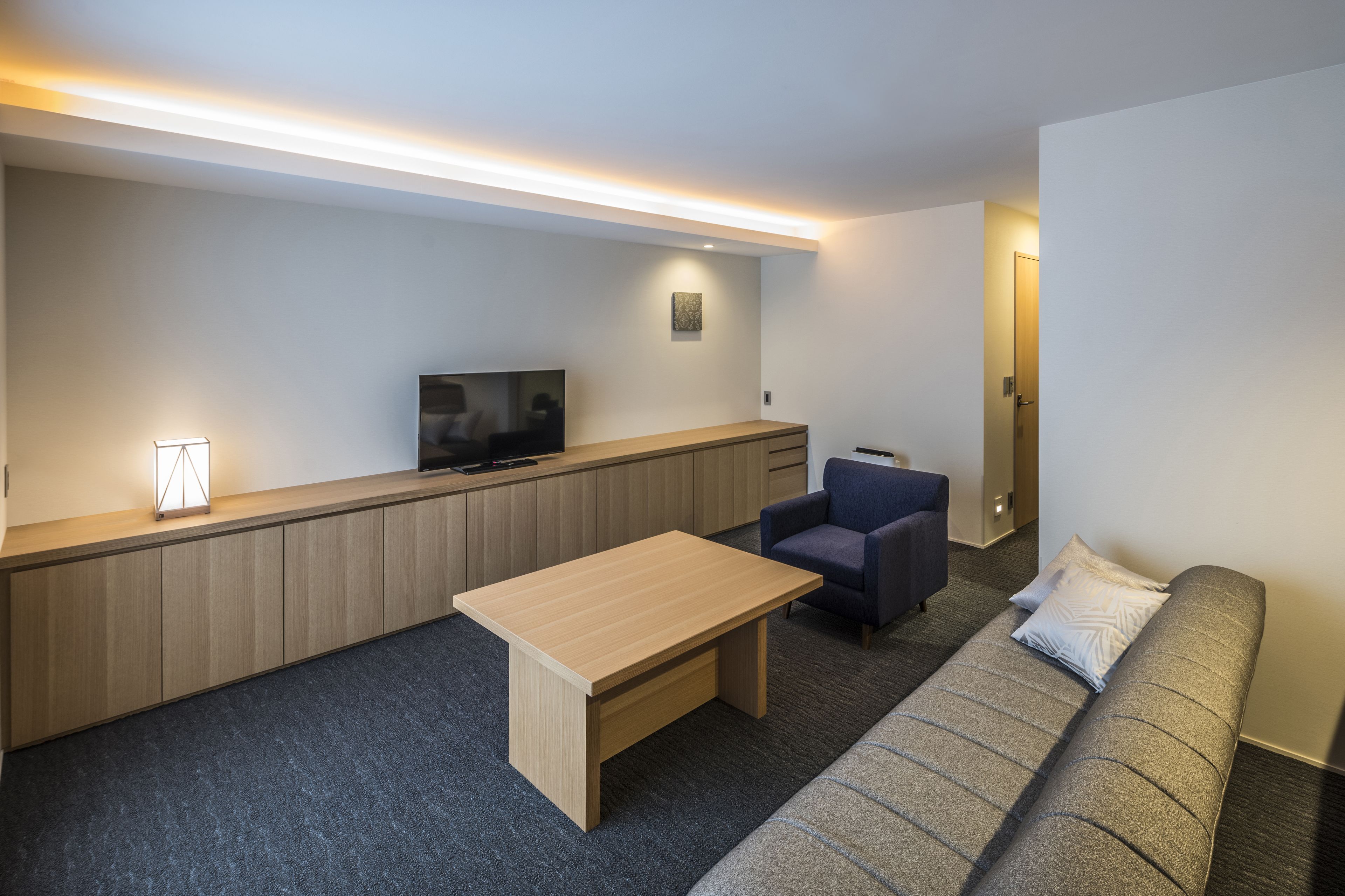 A modern hotel room with a flat-screen TV, a wooden desk, a sofa, and a chair.