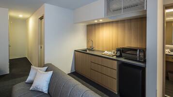 A compact kitchenette with a microwave, coffee maker, and a sink. There is a grey sofa with cushions in the adjacent living area.