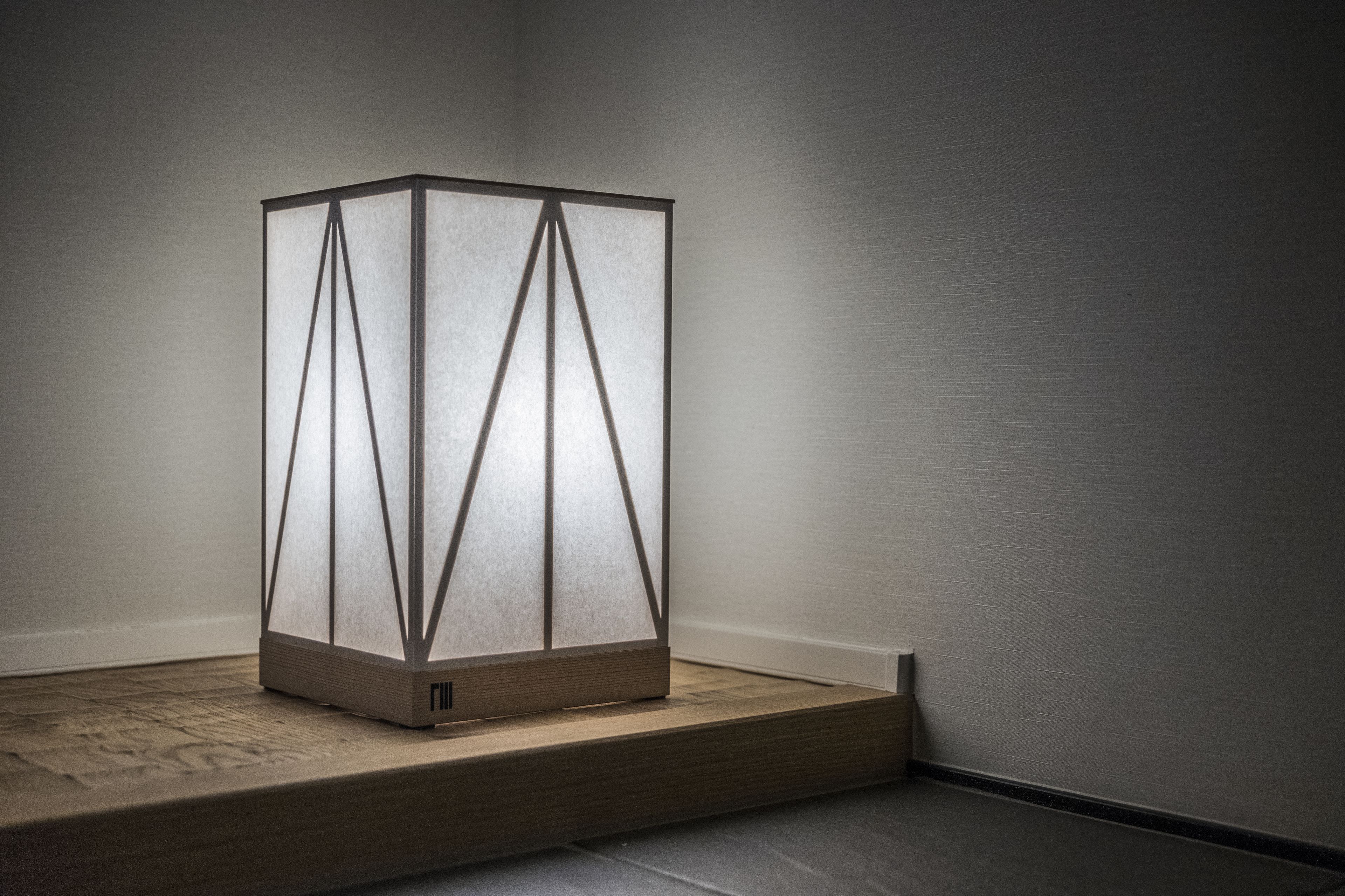 A modern, minimalist floor lamp with a geometric design, placed on a wooden platform against a plain wall.