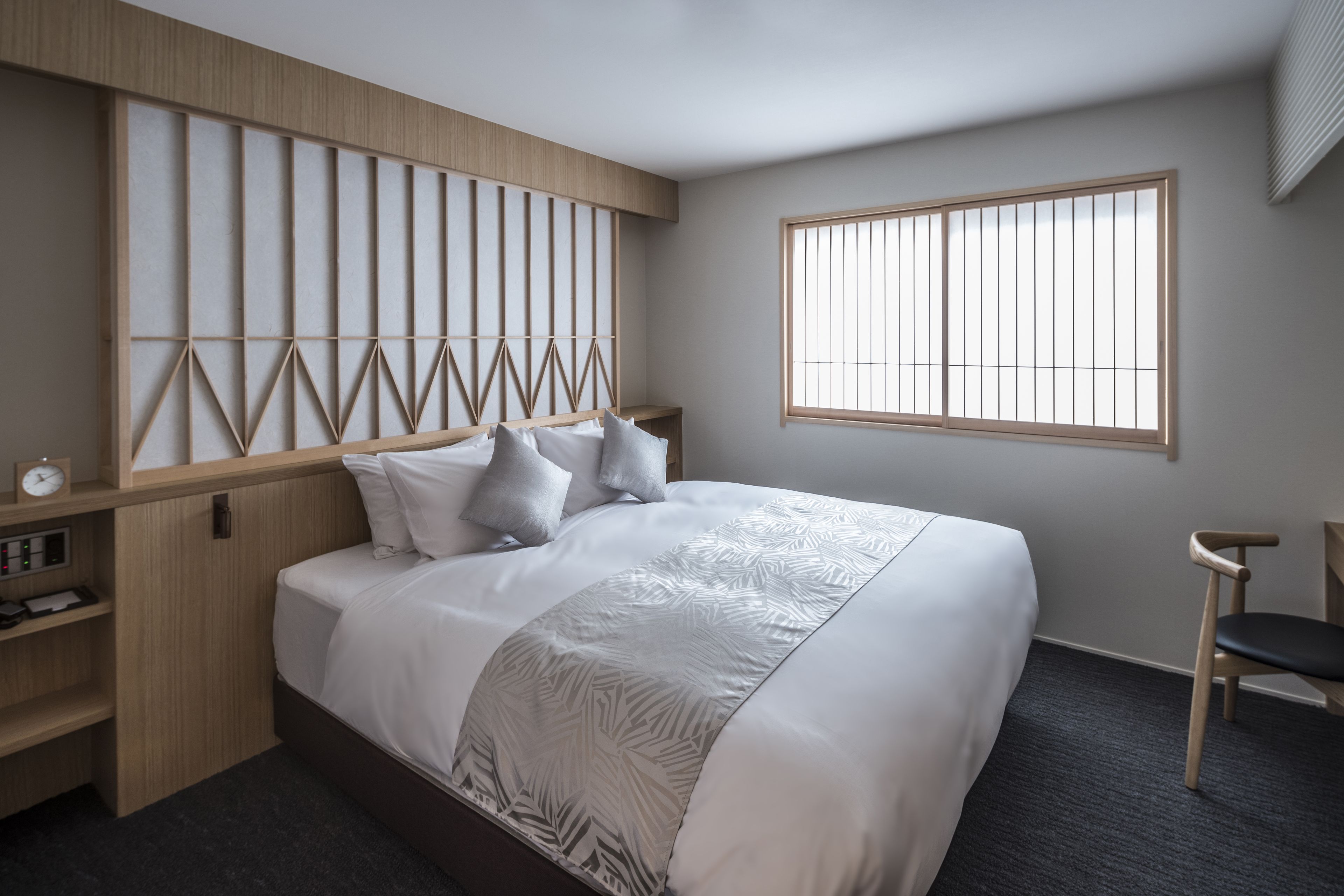A modern hotel room with a large bed, a wooden headboard, a window, a chair, and a small desk with a clock.