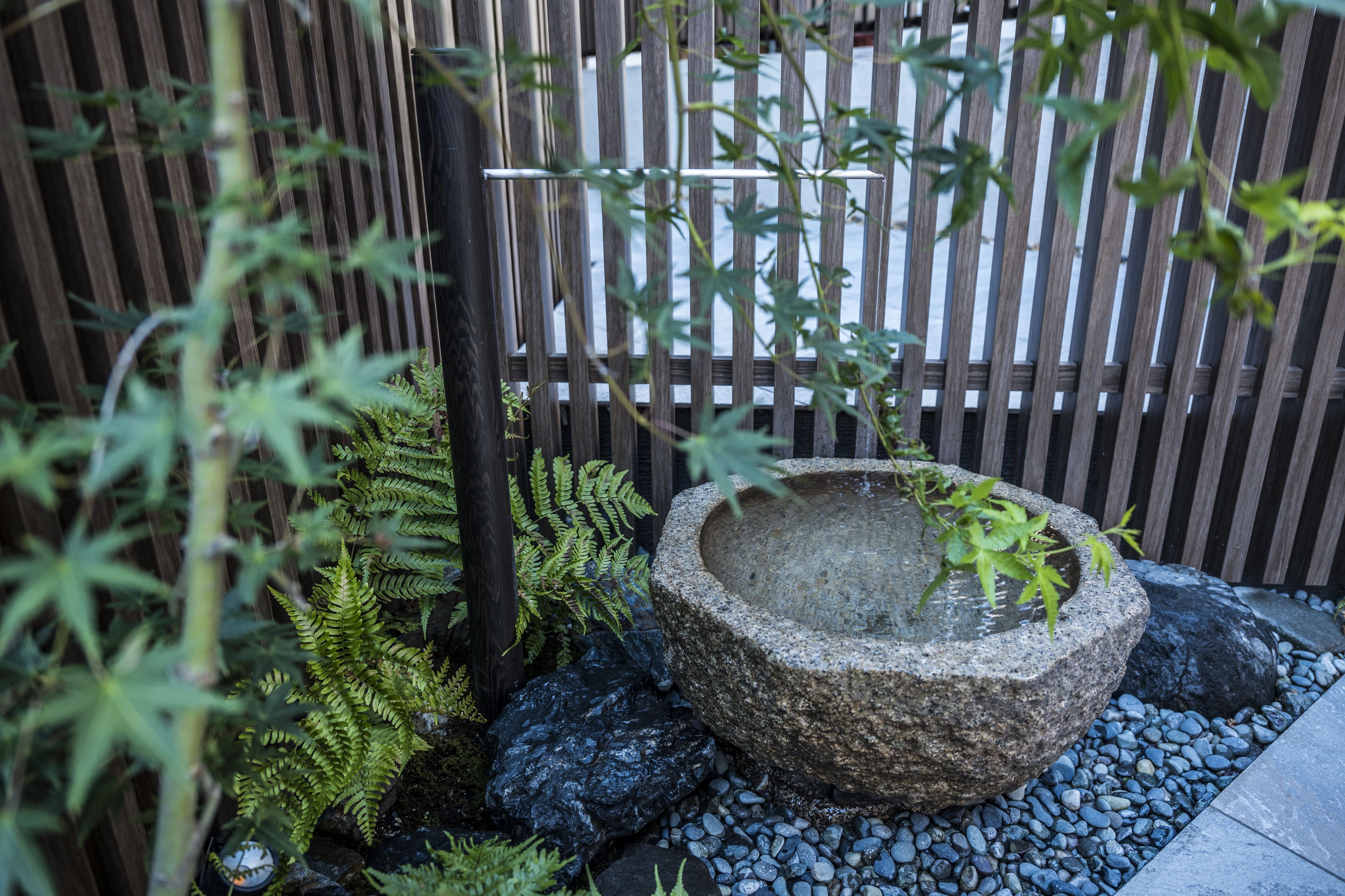 A stone water feature surrounded by rocks and ferns, with a bamboo fence in the background.