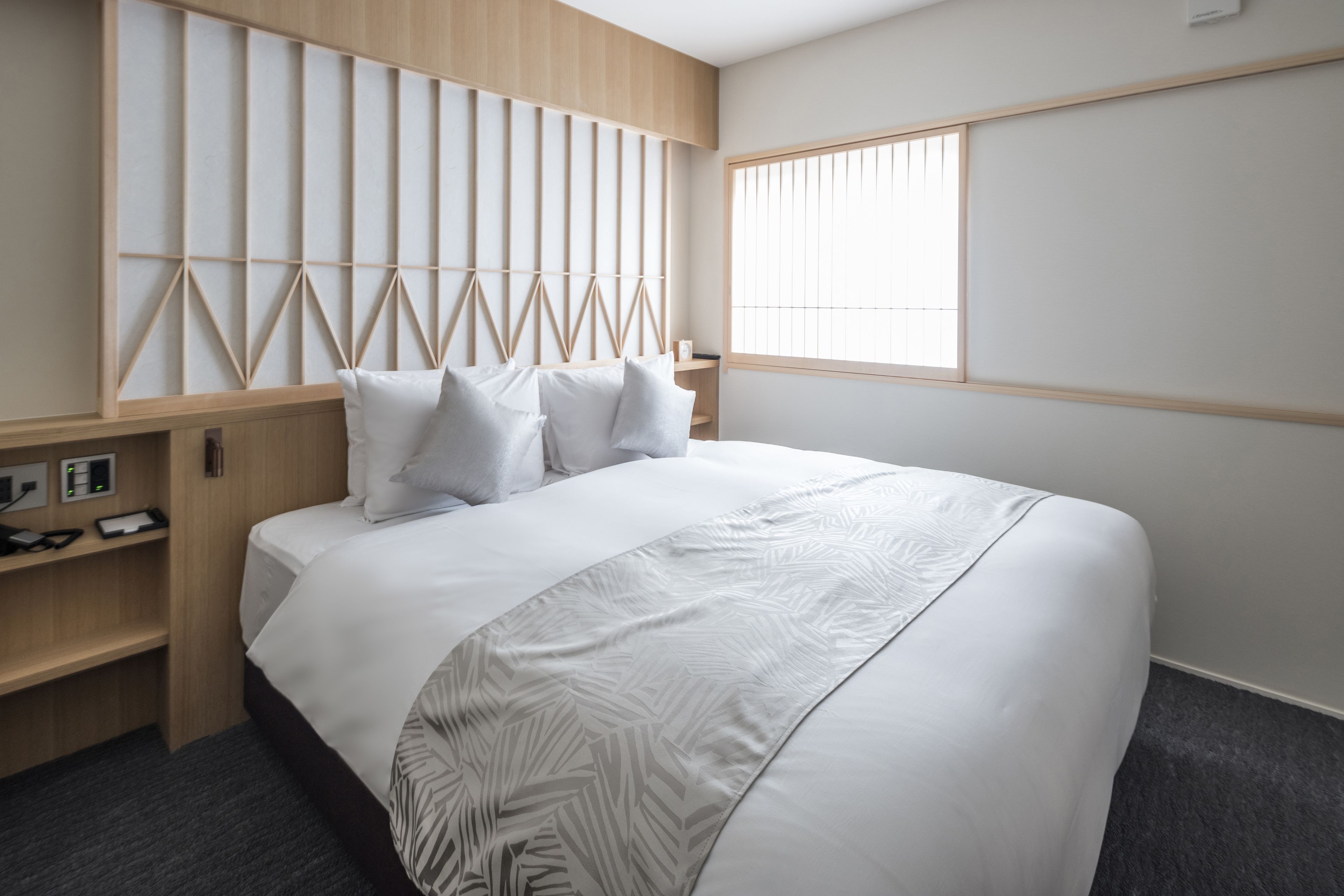 A modern hotel room with a large bed, a wooden headboard, a window with blinds, and a small bedside table with a lamp.