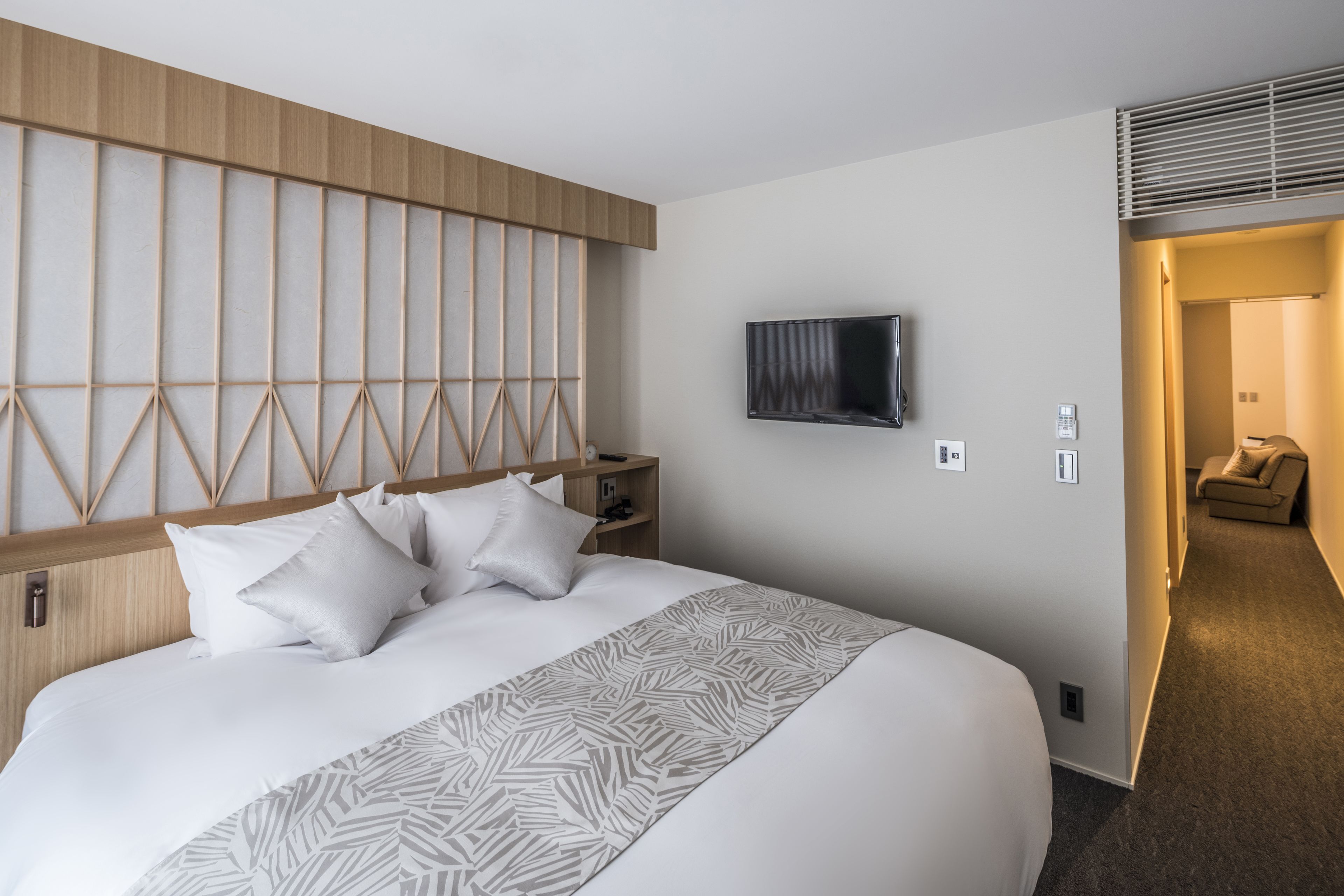 A hotel room with a large bed, a TV mounted on the wall, a wooden headboard, and a small seating area with a sofa.