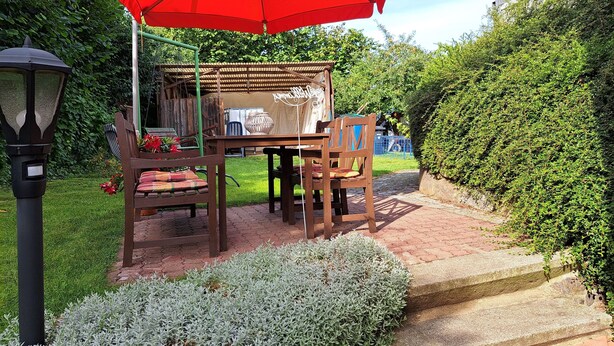 An outdoor seating area with a table and chairs under a red umbrella, surrounded by greenery and a lamp post. - Sylvana vacation apartment (Klein Vielen)