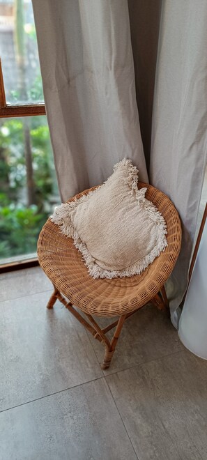 A wicker chair with a textured cushion, positioned near a window with curtains, on a tiled floor.