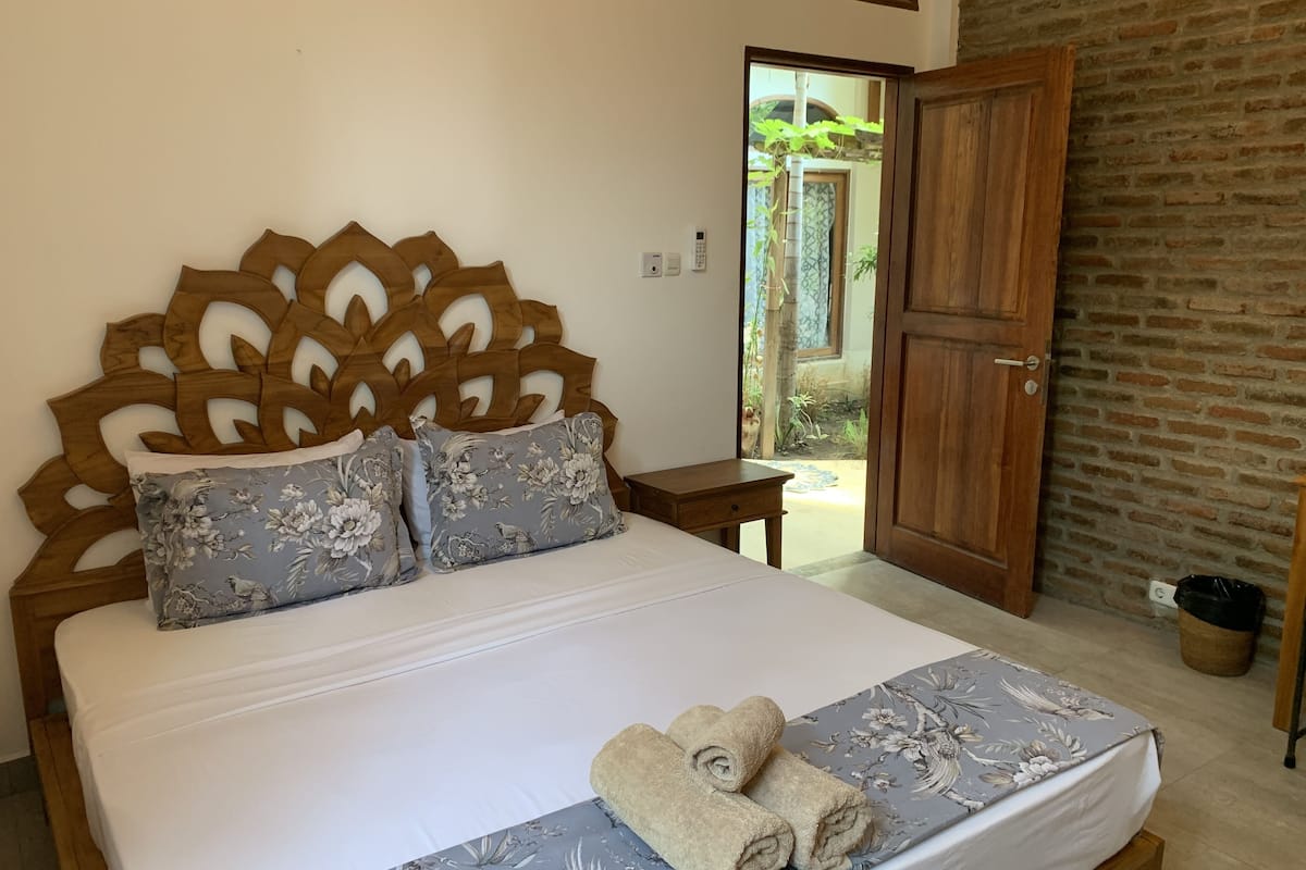 A neatly made bed with floral-patterned pillows and a folded towel, a wooden headboard, a small bedside table, and a view of a brick wall and door leading outside.