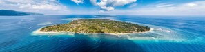 An aerial view of a tropical island with clear turquoise waters, surrounded by lush greenery and a few scattered buildings.