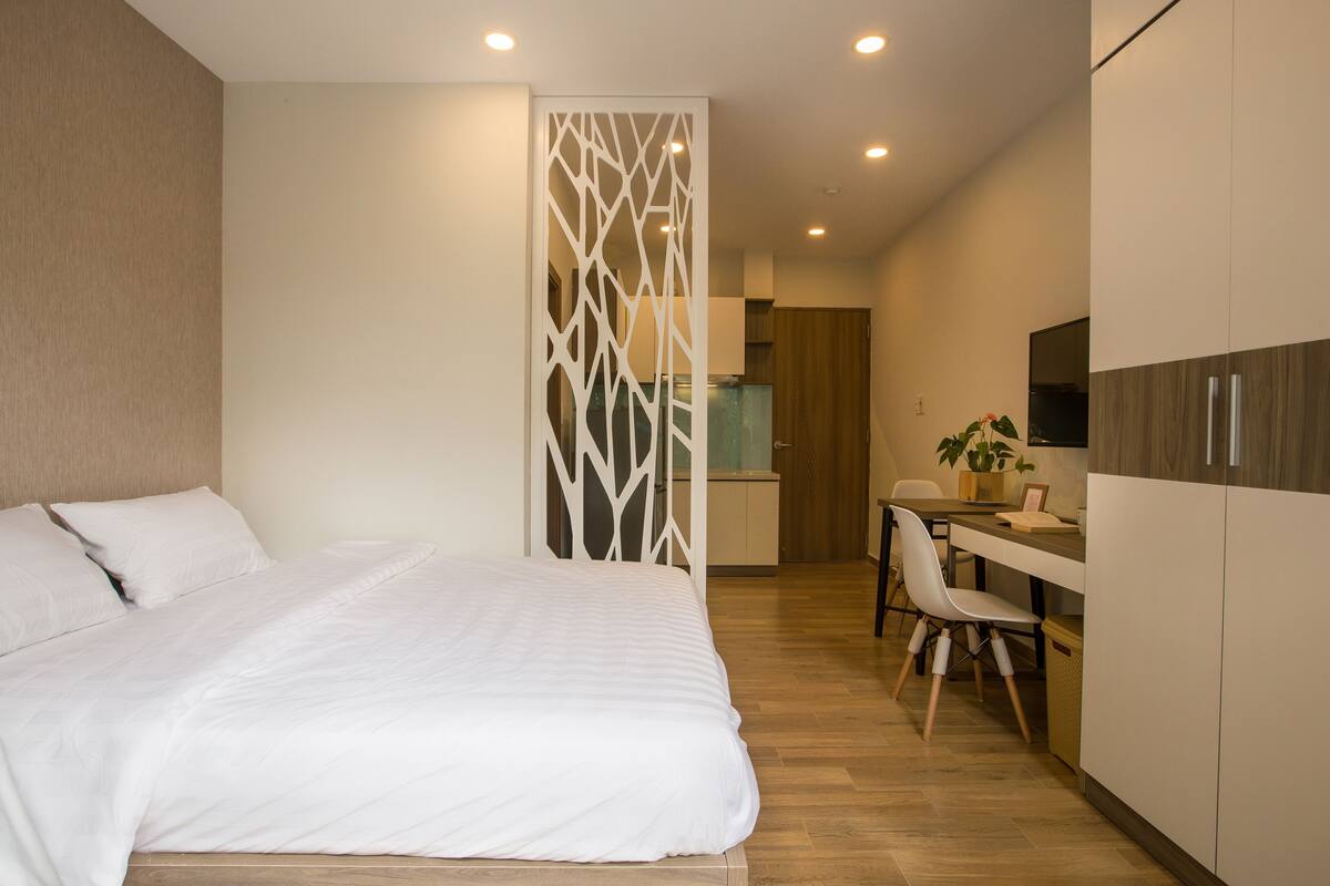 Een moderne hotelkamer met een groot bed, een bureau met stoel, een kitchenette en een decoratieve scheidingswand.