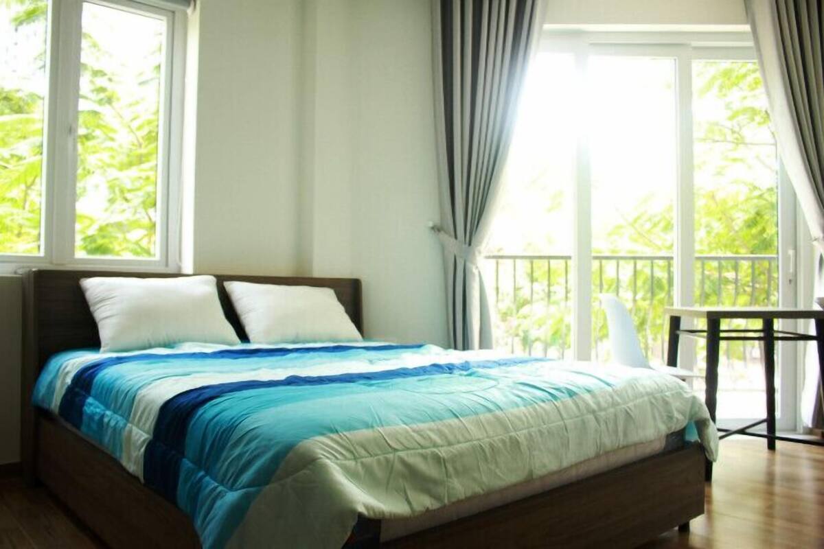 Een bed met een blauw-wit gestreepte spreideken, een houten hoofdbord en uitzicht op groen buiten door grote ramen.