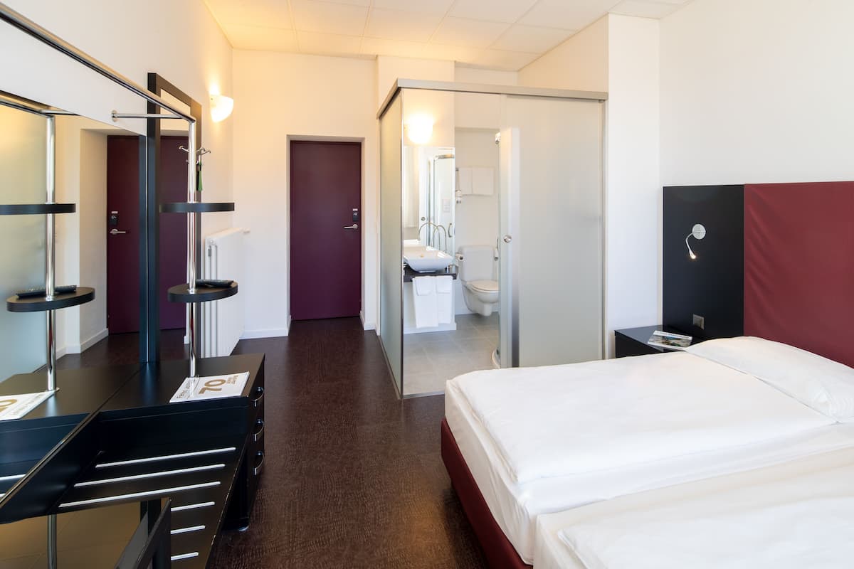 A hotel room with a bed, a desk, a mirror, a wardrobe, and a bathroom visible through an open door.