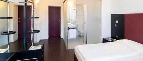 A hotel room with a bed, a desk, a mirror, a wardrobe, and a bathroom visible through an open door.